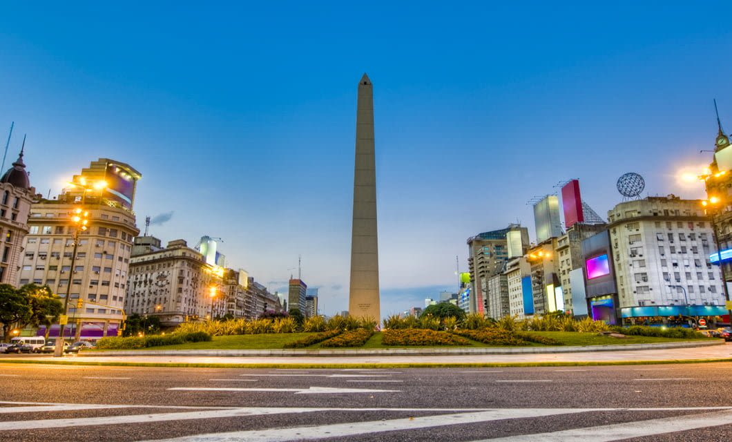 The photo shows the Obelisk of Buenos Aires, a tall stone monument in the center of a city plaza, surrounded by buildings and streetlights during early evening—a must-see for anyone beginning their South American cruise from Buenos Aires.