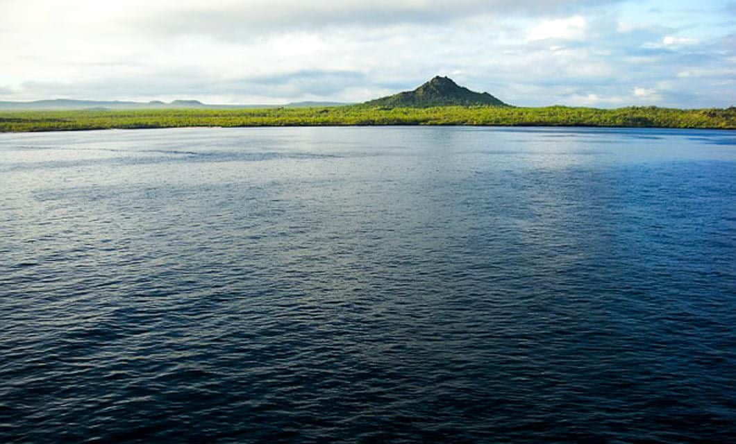 A calm, deep blue sea stretches toward a distant, green island with a single dark volcanic hill under a partly cloudy sky—an inviting scene often witnessed on a Galapagos Islands cruise.