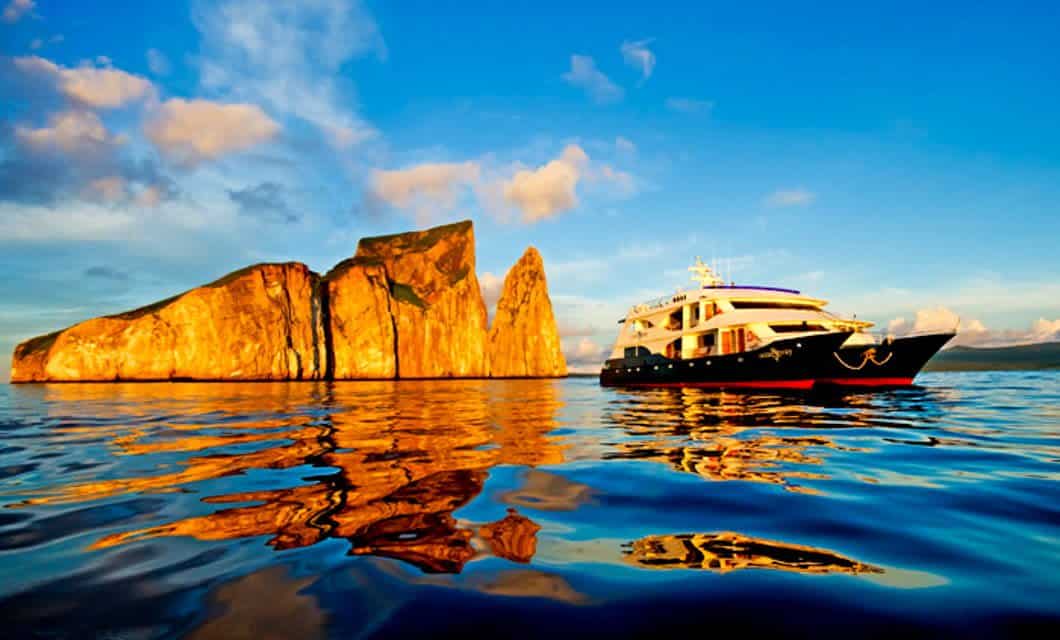 A luxury yacht from the Ocean Spray Galapagos Cruise floats on calm, reflective blue water near a striking rocky formation at sunset, with golden light illuminating the rocks and clouds in the sky.