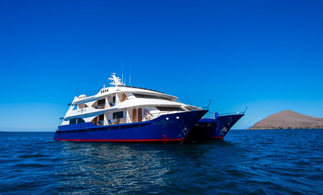 A large, modern blue and white yacht from the Ocean Spray Galapagos Cruise floats on calm blue water under a clear sky, with a rocky island visible in the background.