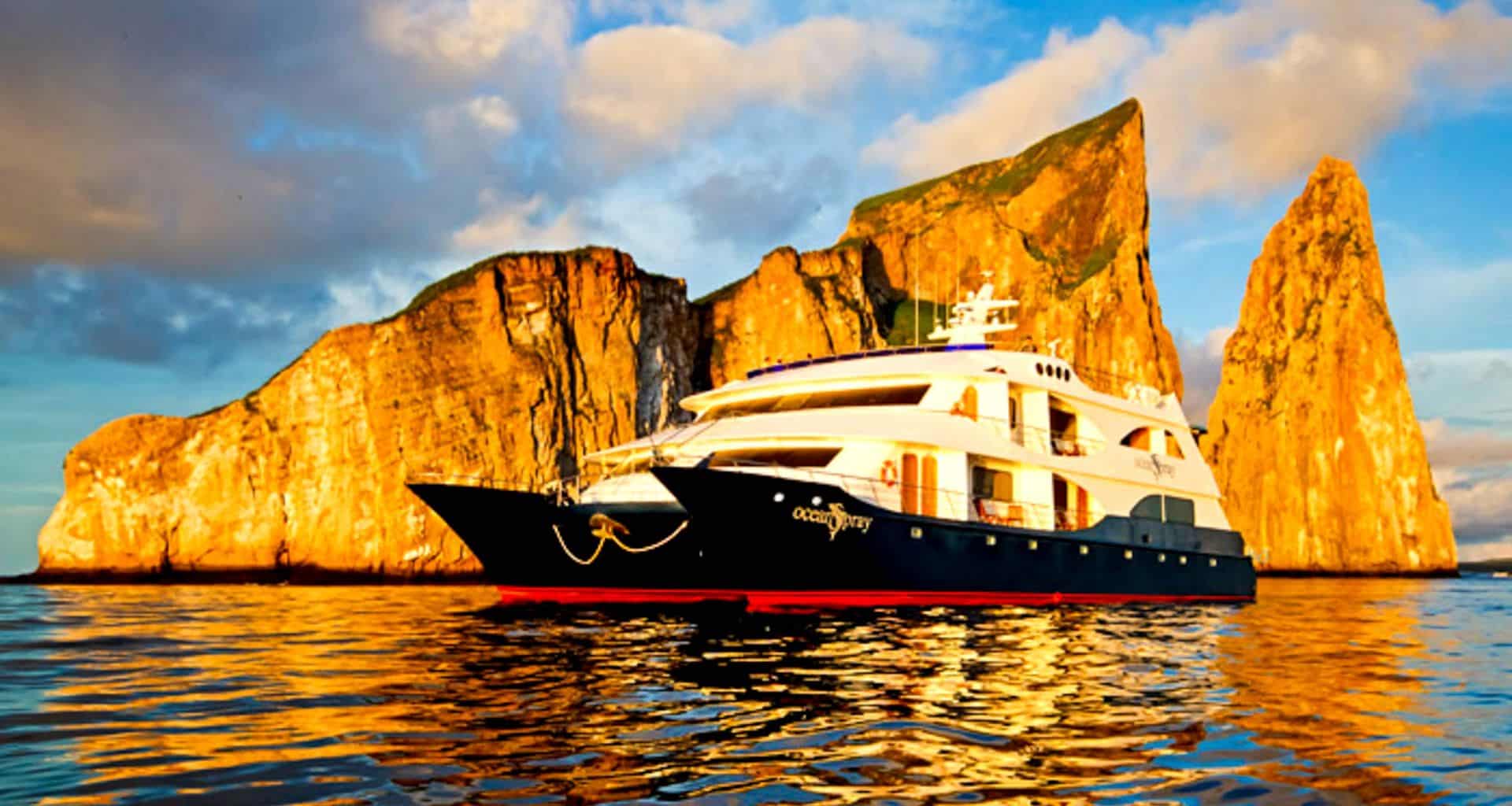 A large white and blue Ocean Spray Galapagos Cruise yacht floats on calm golden water near striking rocky cliffs under a partly cloudy sky at sunset.