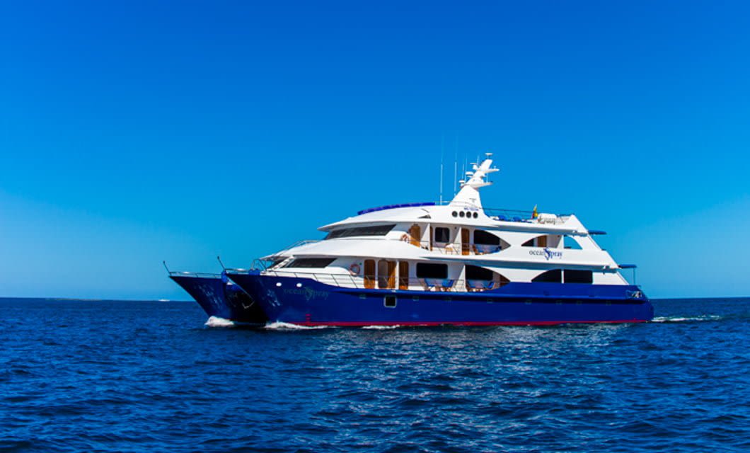 A large, modern blue and white yacht from the Ocean Spray Galapagos Cruise sails on a calm ocean under a clear blue sky.
