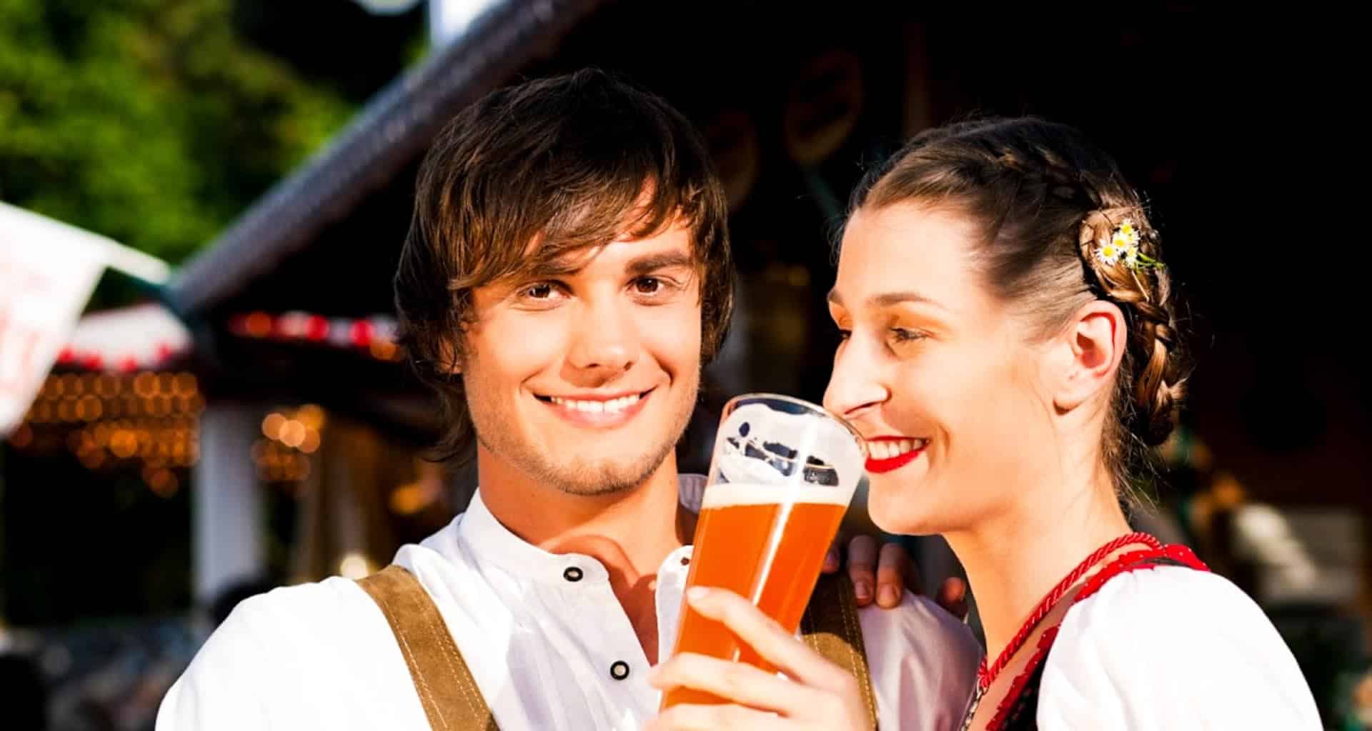 A smiling man and woman in traditional Bavarian clothing enjoy a glass of beer outdoors during a lively Brazil Oktoberfest, with festive decorations blurred in the background.