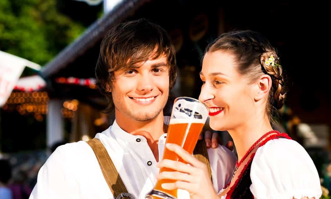 A smiling man and woman in traditional Bavarian clothing stand close together outdoors at Brazil Oktoberfest. The woman holds a tall glass of beer and smiles, while the man looks at the camera. The blurred background hints at a festive celebration.