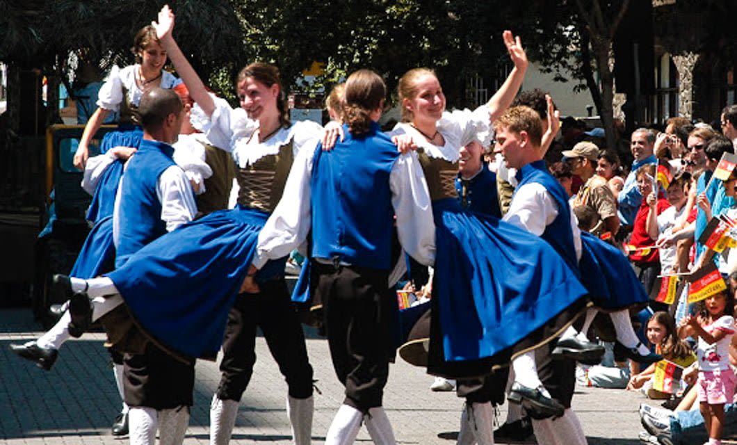 A group of dancers in traditional blue and white folk costumes perform outdoors during a Brazil Oktoberfest, lifting and twirling each other as a crowd waves small flags. The festive scene is lively, set against trees in the background.