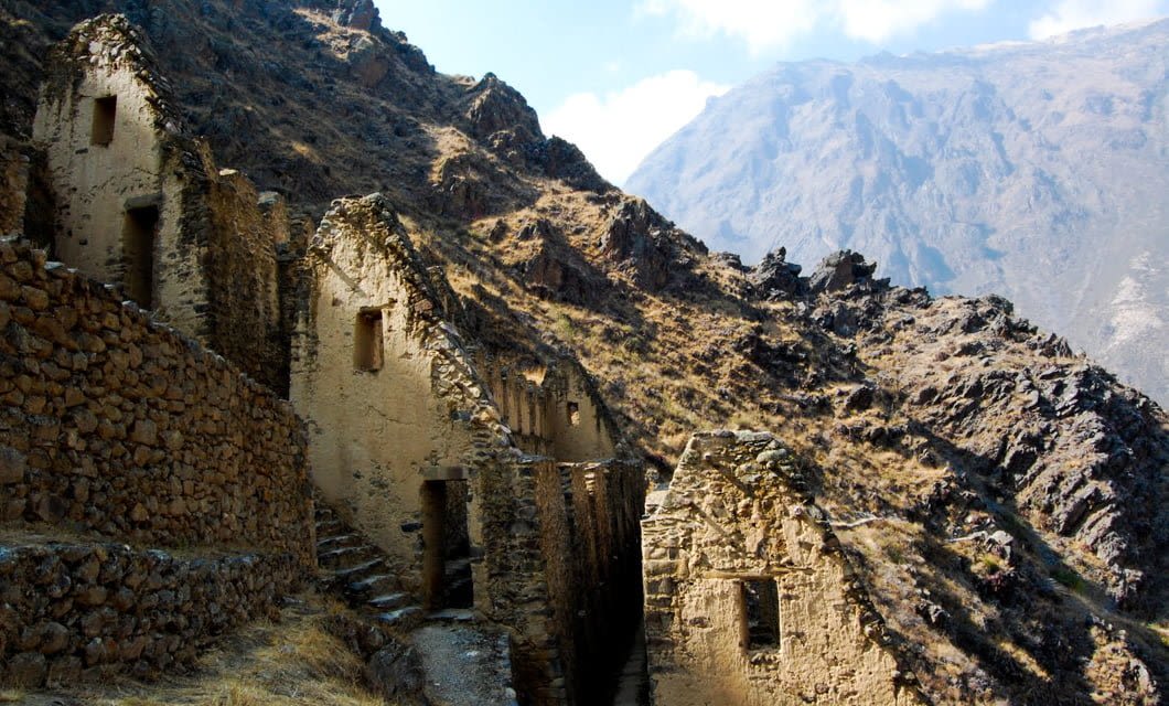 Ancient stone ruins with doorways sit on a steep, rocky mountainside along the Lares Trek, under a partly cloudy sky, with rugged peaks rising in the background.