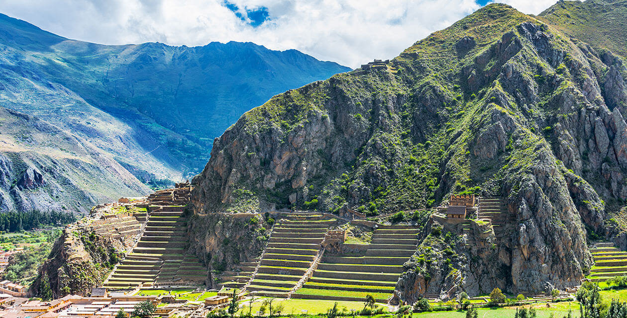 Terraced stone ruins climb a steep, green mountainside under a partly cloudy sky, with rugged peaks and a lush valley in the background—a breathtaking scene among South American Wonders, perfect for an unforgettable Honeymoon South America.
