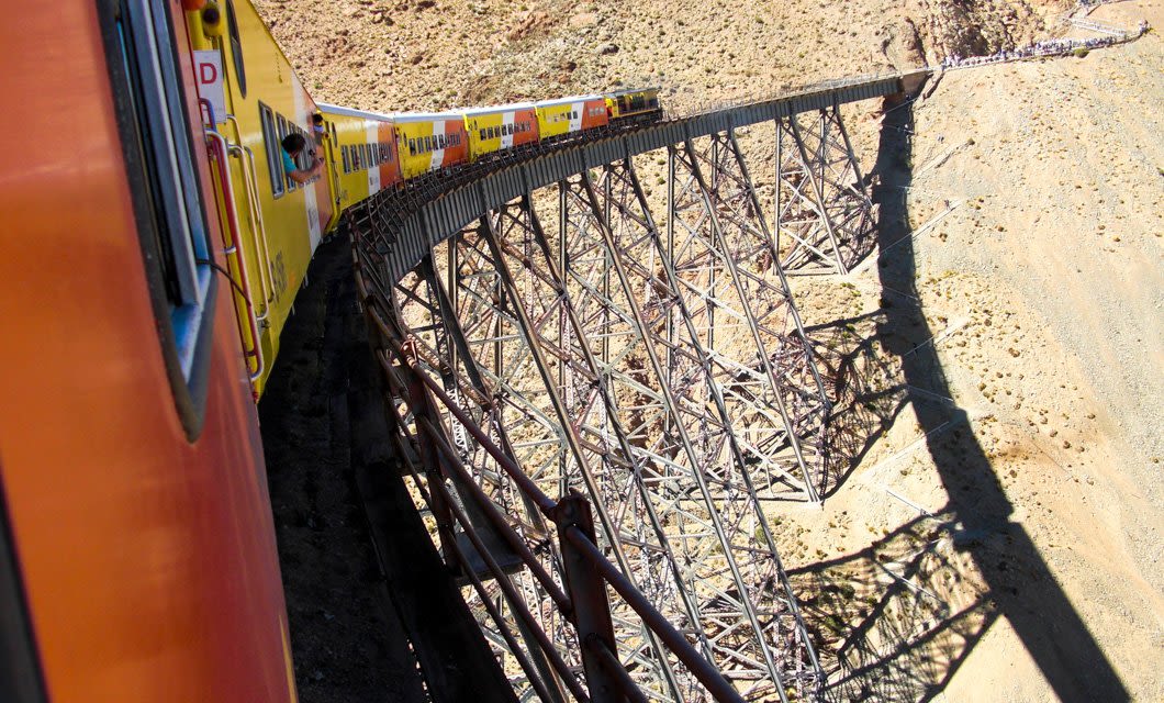 A yellow and orange train crosses a tall, narrow metal bridge over a dry, rocky canyon in the Andes. The bridge’s steel framework casts a shadow on the ground below. The photo is taken from a window of the Magic Triangle route.