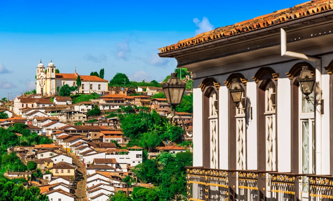 A scenic view of a hillside town with colonial-style houses, a white church with twin towers, lush greenery, and a close-up of a historic building’s balcony—showcasing Colonial Treasures—under a bright blue sky.