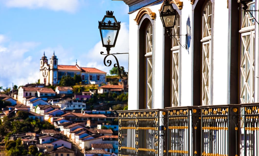 A close-up of a colonial-style balcony and lamp with a view of colorful houses and a historic church on a hillside along the Brazil Gold Route, all beneath a vibrant blue sky.