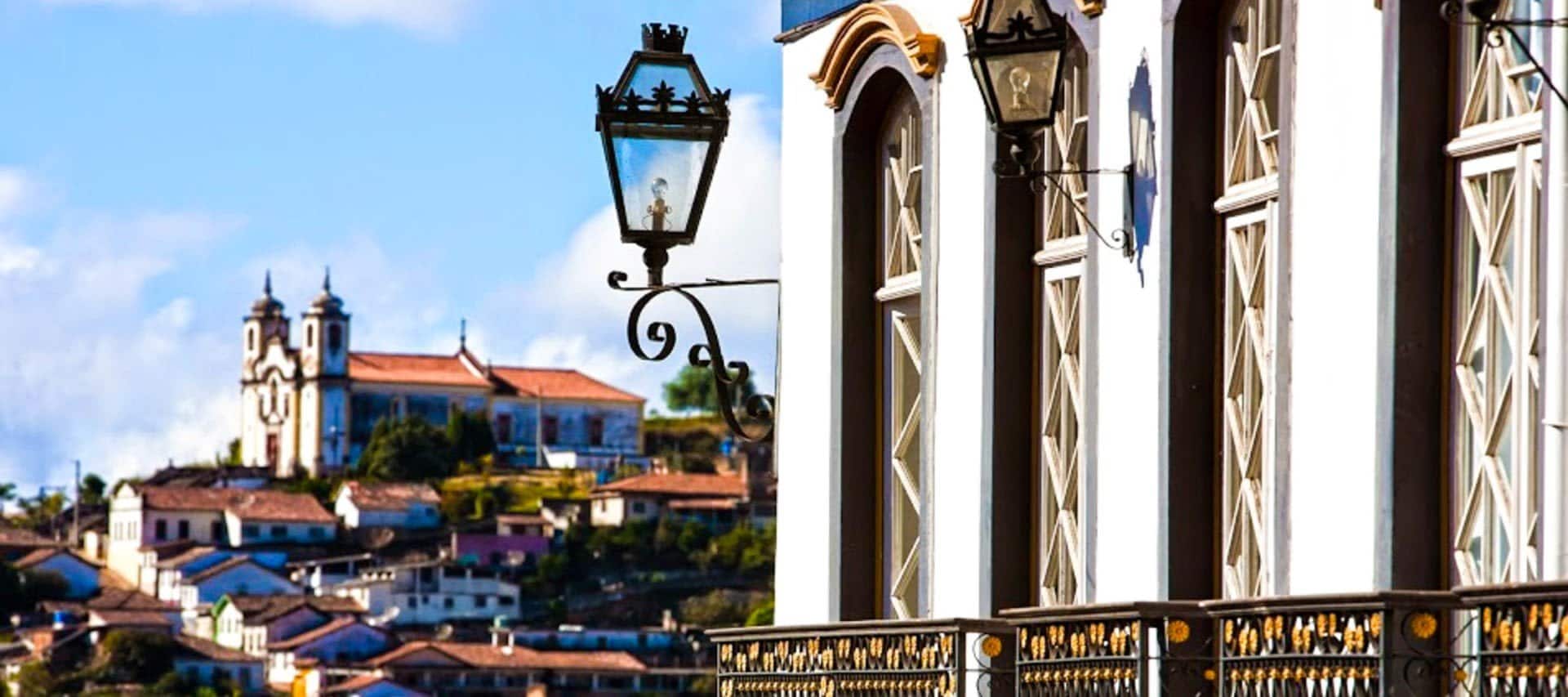 A close-up view of ornate street lamps and windows on a colonial building in Ouro Preto, with a hillside town and a large church in the background under a blue sky—perfect for those interested in Ouro Preto travel.