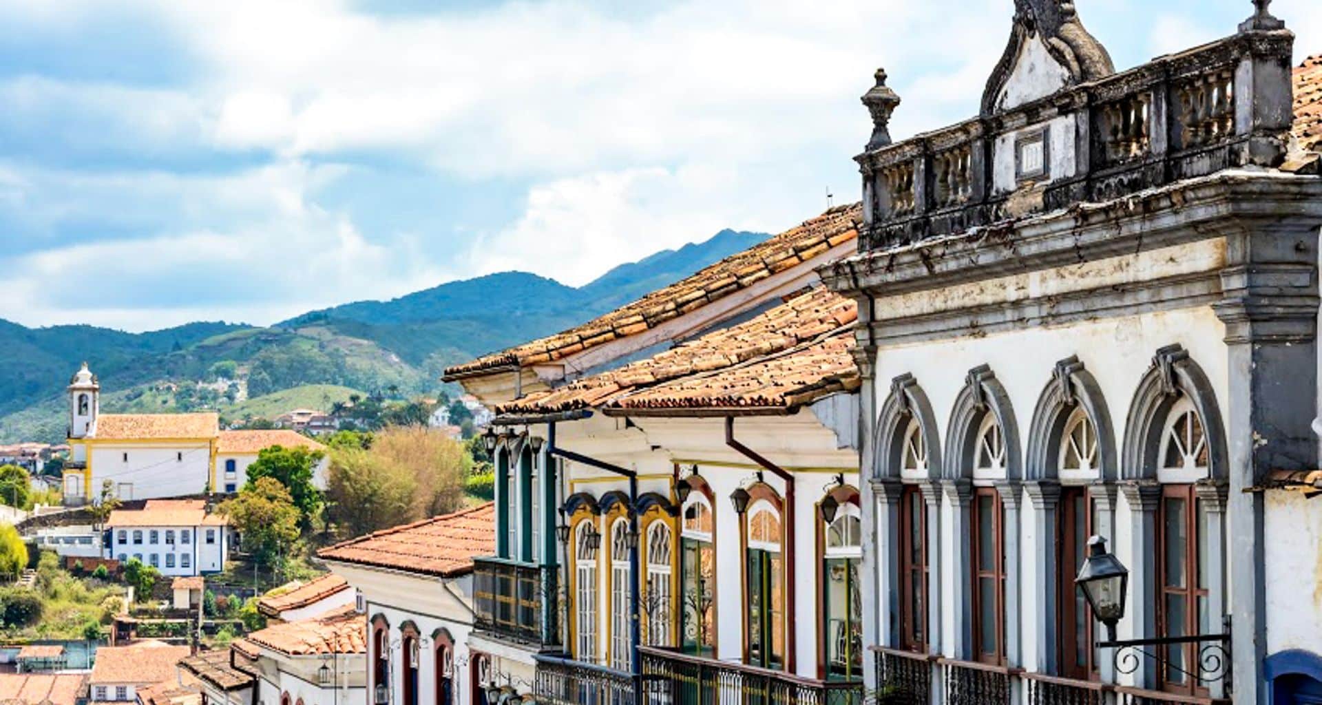 Colorful colonial-style buildings with tiled roofs line a street in the historic town of Ouro Preto, set against green hills and a cloudy sky. A church with a bell tower rises in the distance, capturing the charm of this Brazil Amazon Tour destination.