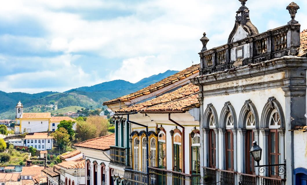 Colorful colonial-style buildings with ornate balconies line a street in the hilly town of Ouro Preto, with a church and lush green mountains in the background under a partly cloudy sky.