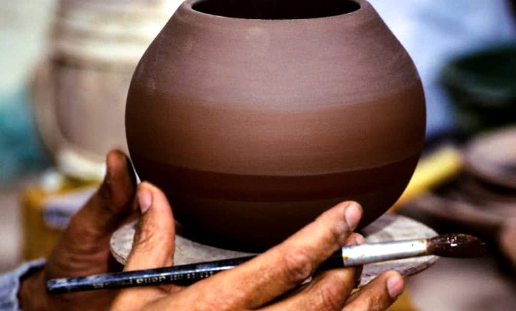A person holds a round, unglazed clay pot while applying paint or glaze to it with a brush, capturing the artistry often admired on a Peru train tour and evoking the charm of handmade pottery seen along a Peru Bolivia rail journey.