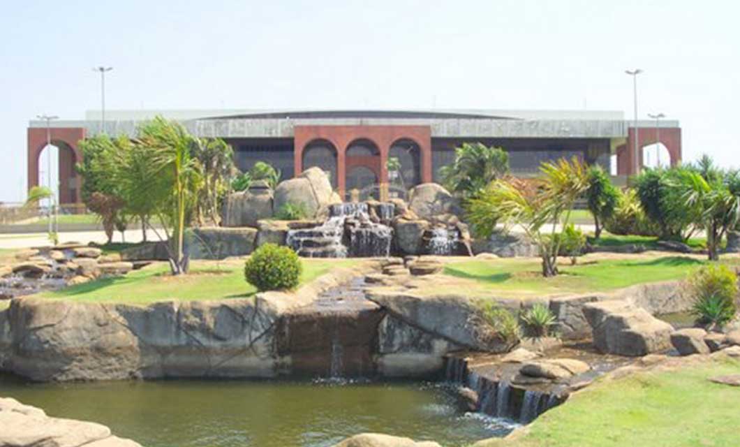 A landscaped garden inspired by Brazil's Cerrado features a small waterfall and pond, lush greenery, and a large building with red arches and glass windows. Perfect for a tranquil tour under a clear, bright sky.