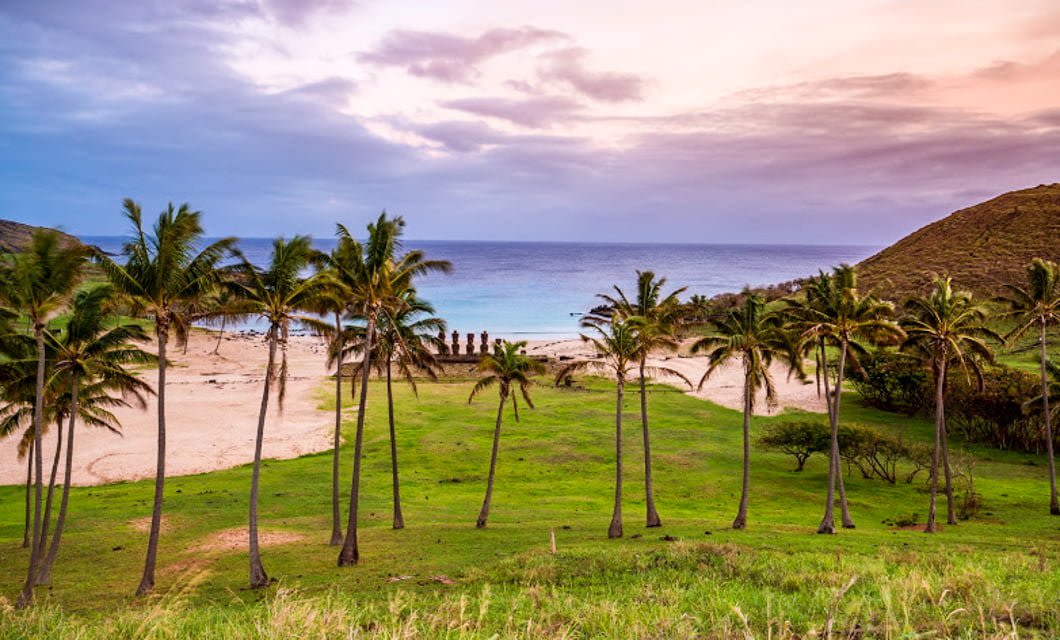 Palm trees on a grassy hill overlook a sandy beach with moai statues on Easter Island, reminiscent of the ancient wonders found at Machu Picchu, all set against a colorful sunset sky and a tranquil ocean backdrop.