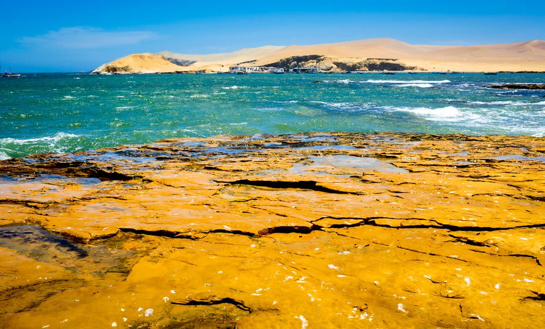 Rocky yellow coastline with cracks in the foreground, blue-green ocean waves in the middle, and sandy desert dunes under a clear blue sky capture the essence of Peru’s dramatic landscapes.