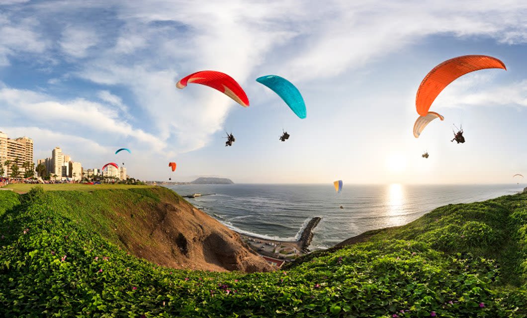 Paragliders with colorful parachutes soar above a green cliff overlooking the ocean at sunset, near city buildings, with views reminiscent of Easter Island and a clear sky in the background.