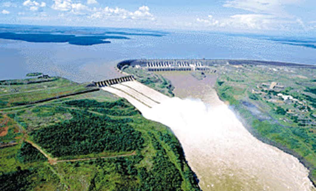 Aerial view of a large hydroelectric dam near Asuncion releasing water into a river, surrounded by lush green vegetation and a wide expanse of water with the Chaco in the background under a partly cloudy sky.