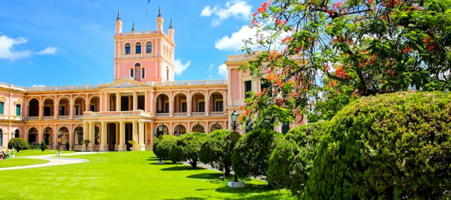 A grand pink and yellow neoclassical building with towers and arches stands under a bright blue sky, surrounded by manicured green lawns—an iconic sight for those exploring Paraguay travel and tours.