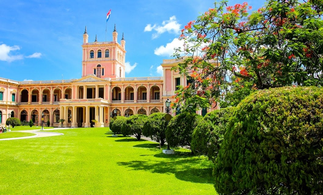 A grand pink historic building with arched windows and towers, echoing the legacy of the Incas, stands behind manicured green lawns and trees under a bright blue sky. The national flag flies atop the central tower.