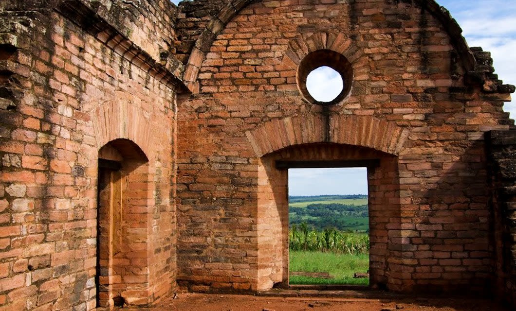 Ruins of an old brick building with arched doorways and a circular window, looking out onto a green landscape under a partly cloudy sky—a hidden gem to discover on your Paraguay Tour near Iguazu.