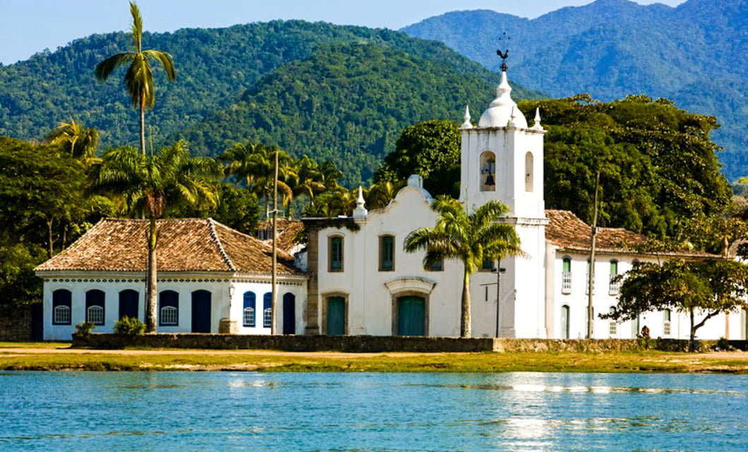 A white colonial-style church with a bell tower and blue-trimmed windows stands by the water, surrounded by palm trees and lush green mountains—an iconic stop on the Brazil Gold Route under a clear sky.