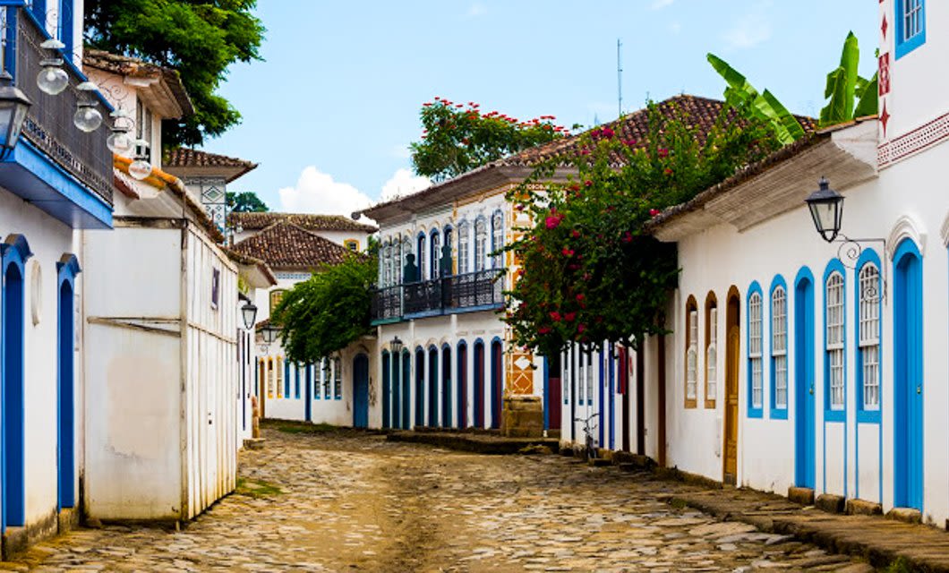 A cobblestone street in Paraty, lined with colorful colonial-style buildings with white walls, blue doors, and flower-covered balconies, evokes the charm of a tropical island under a partly cloudy sky.