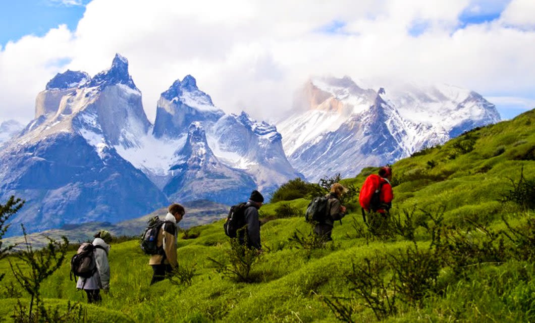 A group of hikers with backpacks walk across green hills, with dramatic snow-capped mountains and a partly cloudy sky in the background—one of the unforgettable Chile highlights for adventure lovers.