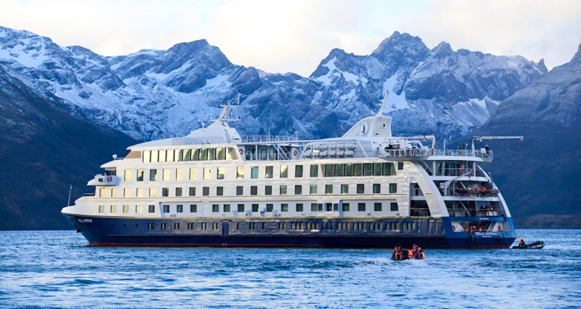 A large white and blue Patagonia Cruise ship floats on calm water with people in small boats nearby, set against a backdrop of snow-capped, rugged mountain peaks near Torres del Paine.