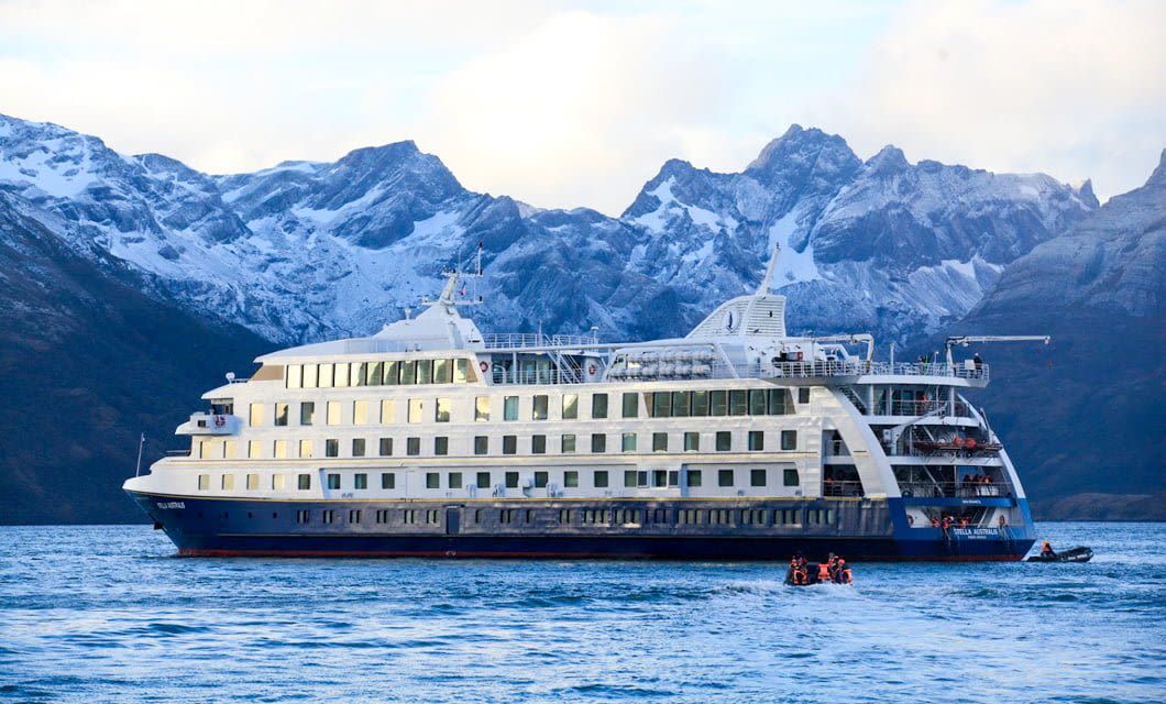 A large white and blue Australis Cruises ship is anchored on calm water in the Fuegian Fjords, with snow-capped mountains of Patagonia in the background. A small boat with people in orange life vests is near the ship.