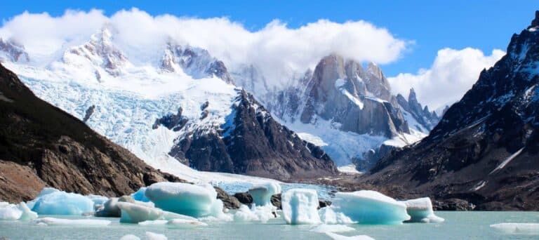 Icebergs float in a turquoise lake surrounded by snow-capped mountains and glaciers under a partly cloudy blue sky. Rocky cliffs frame the scene, creating a breathtaking vista perfect for Patagonia tours.