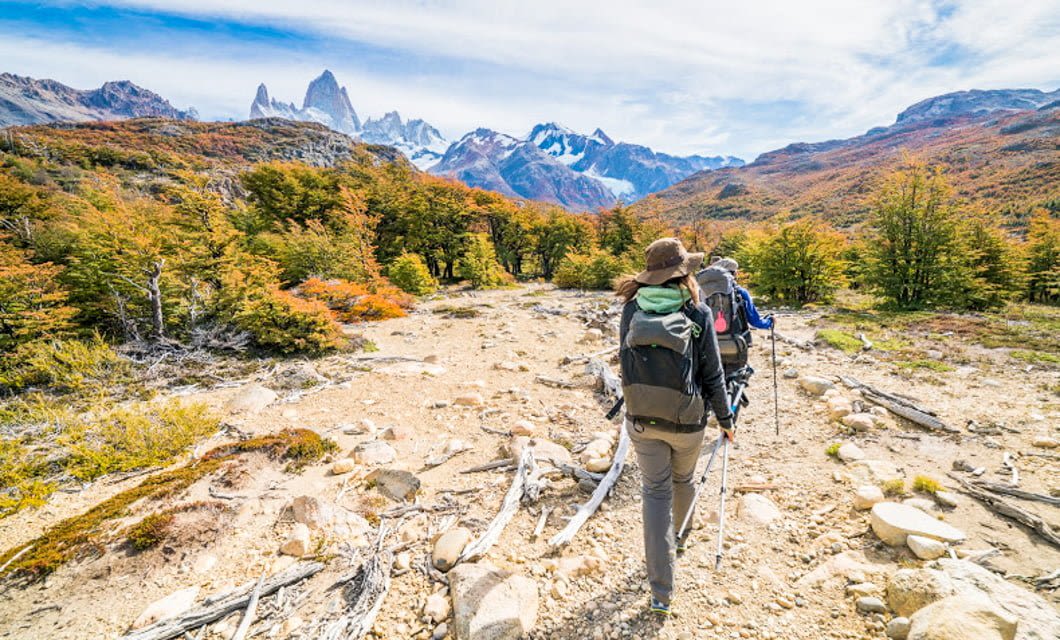 Two hikers with backpacks and trekking poles walk along a rocky trail through colorful autumn trees in Patagonia, with snow-capped mountains and distant glaciers beneath a partly cloudy sky.