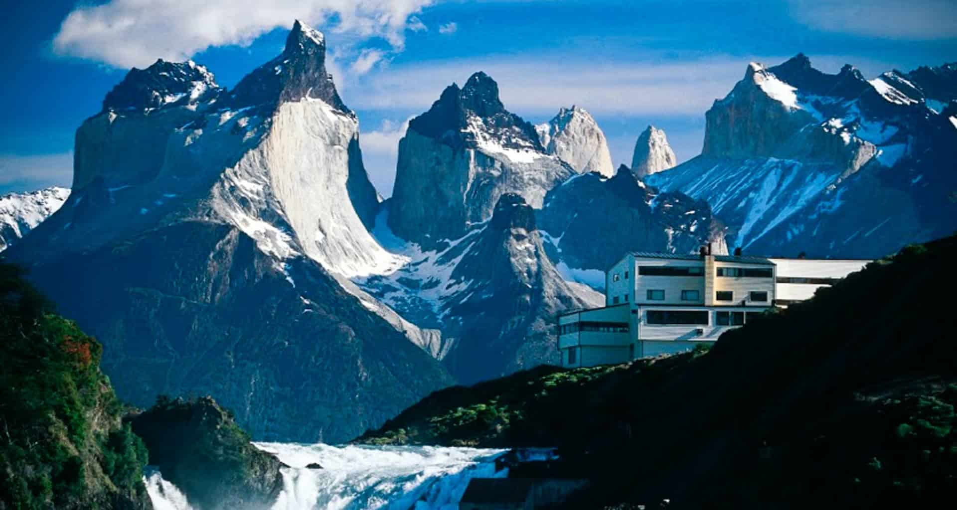 A modern white building, possibly Explora Patagonia, sits near a cascading waterfall with dramatic jagged mountain peaks rising in the background under a blue Patagonia sky with scattered clouds.