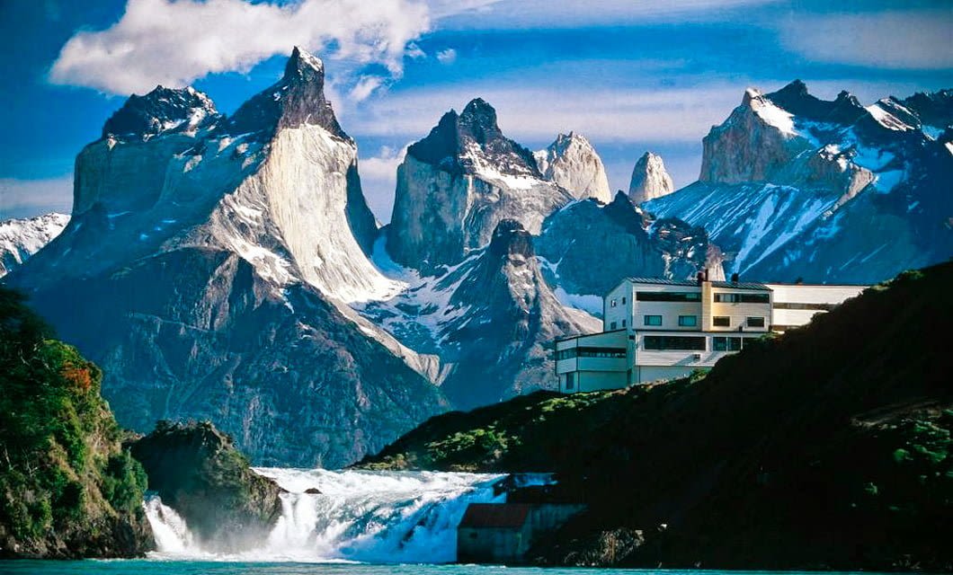 A modern white building sits by a cascading waterfall in Patagonia, with dramatic snow-capped mountain peaks rising sharply in the background under a blue sky.