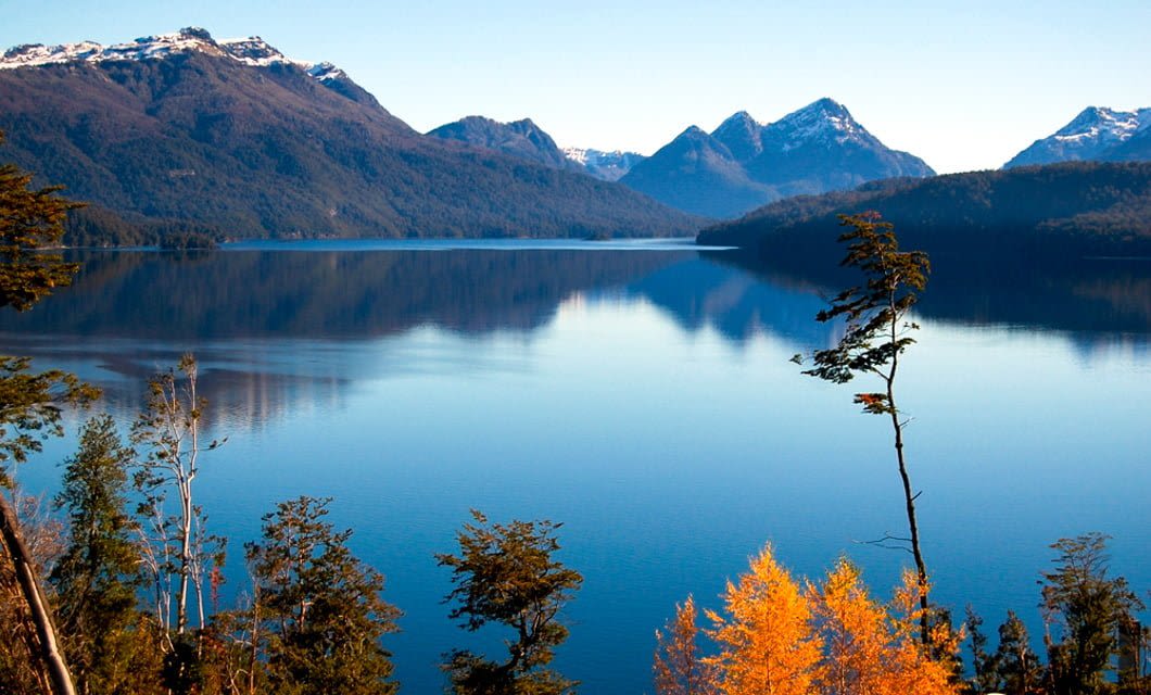 A calm lake reflects surrounding forested mountains with snow-capped peaks under a clear blue sky. Trees with autumn foliage in the foreground add bright yellow and orange to this serene landscape, reminiscent of scenes along a Patagonia Train tour.