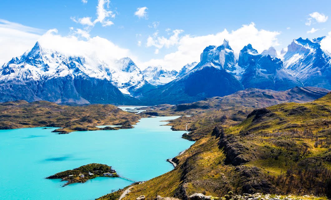 A bright turquoise lake winds through a rugged valley in Southern Patagonia, surrounded by snow-capped mountains under a blue sky with scattered clouds in this dramatic, natural landscape.