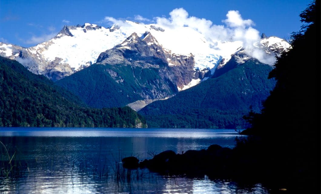 Snow-capped mountains rise above dense green forests, reflected in a calm, clear Patagonian lake under a bright blue sky with scattered clouds. Rocks and reeds are visible in the lake’s foreground.