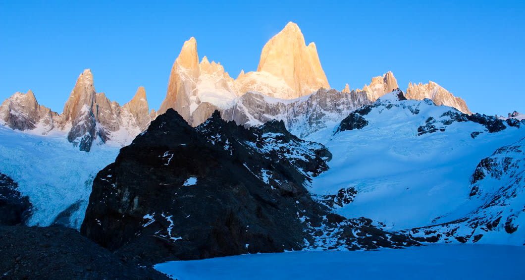 Dramatic mountain peaks, including a tall sunlit summit, rise above rugged dark rocks and snowy slopes under a clear blue sky—an iconic scene from an El Chalten trekking tour near El Calafate.