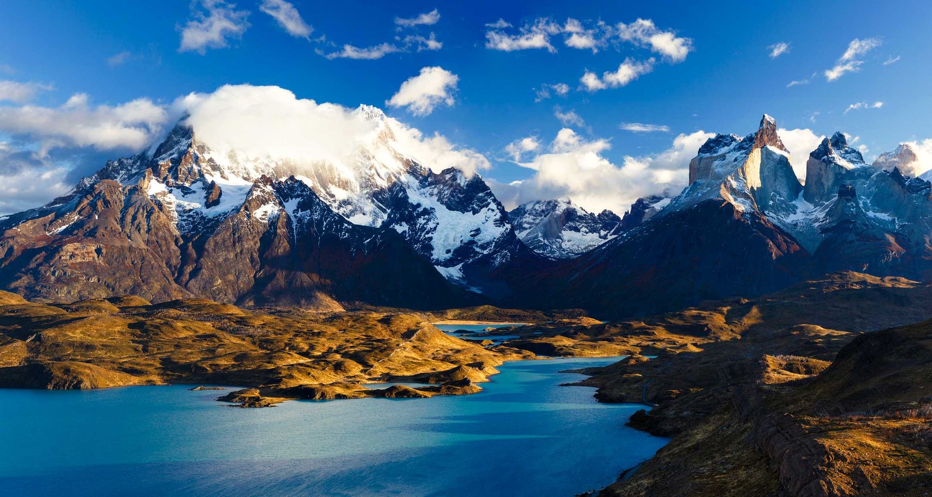 Snow-capped mountains with dramatic peaks rise above turquoise lakes and rugged, golden terrain in Torres del Paine under a bright blue sky with scattered clouds—a breathtaking view on any tour.