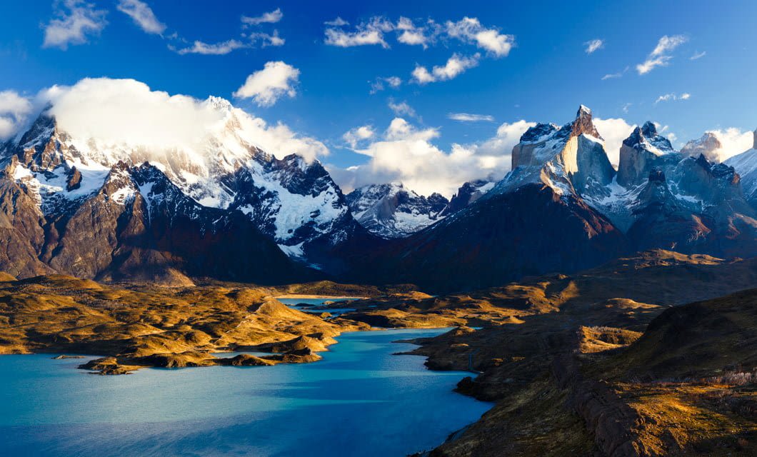 Snow-capped mountains rise behind turquoise lakes and rolling hills under a partly cloudy blue sky, creating a dramatic, sunlit Patagonian landscape—perfect for exploring on a Mare Australis Patagonia Cruise from Santiago to Buenos Aires.