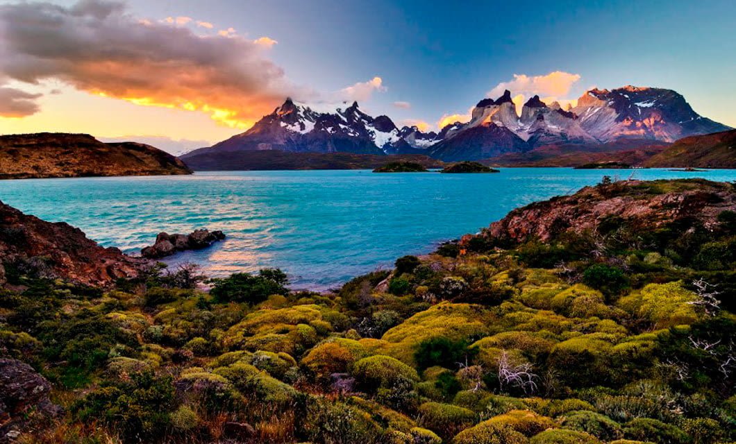 A turquoise lake surrounded by lush green vegetation, with dramatic snow-capped mountains in the background and a vibrant sunset sky—one of the unforgettable Chile highlights for any Chile tourism adventure.