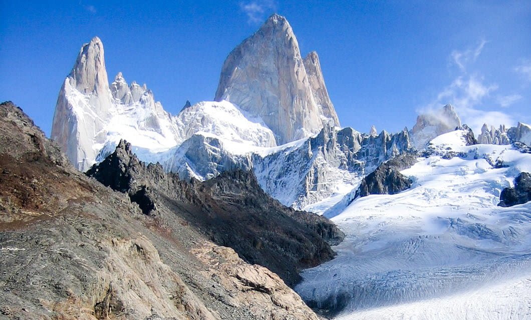 Snow-covered jagged mountains rise sharply under a clear blue sky, with rocky and icy slopes in the foreground. The tallest peak stands prominently at the center, surrounded by smaller peaks and glaciers of wild Patagonia.