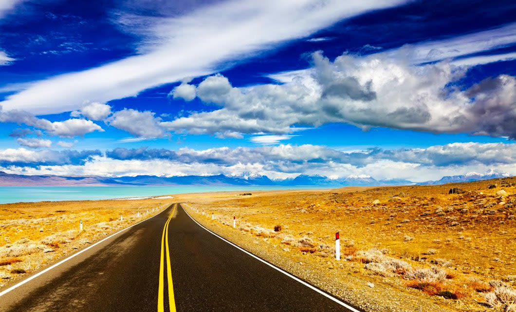 A straight, empty road stretches into the distance along the Carretera Austral, winding through a barren, golden landscape under a dramatic sky with vibrant clouds and distant snow-capped mountains.