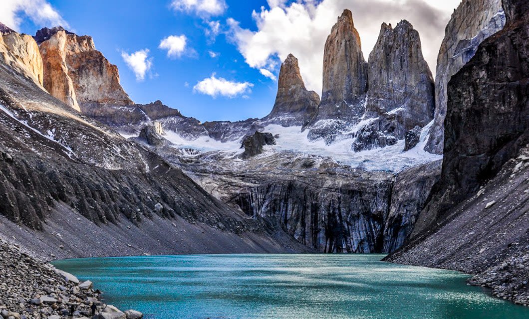 Turquoise glacial lake surrounded by rocky, snow-dusted mountains and jagged peaks under a partly cloudy blue sky—a stunning highlight of Southern Patagonia tourism.