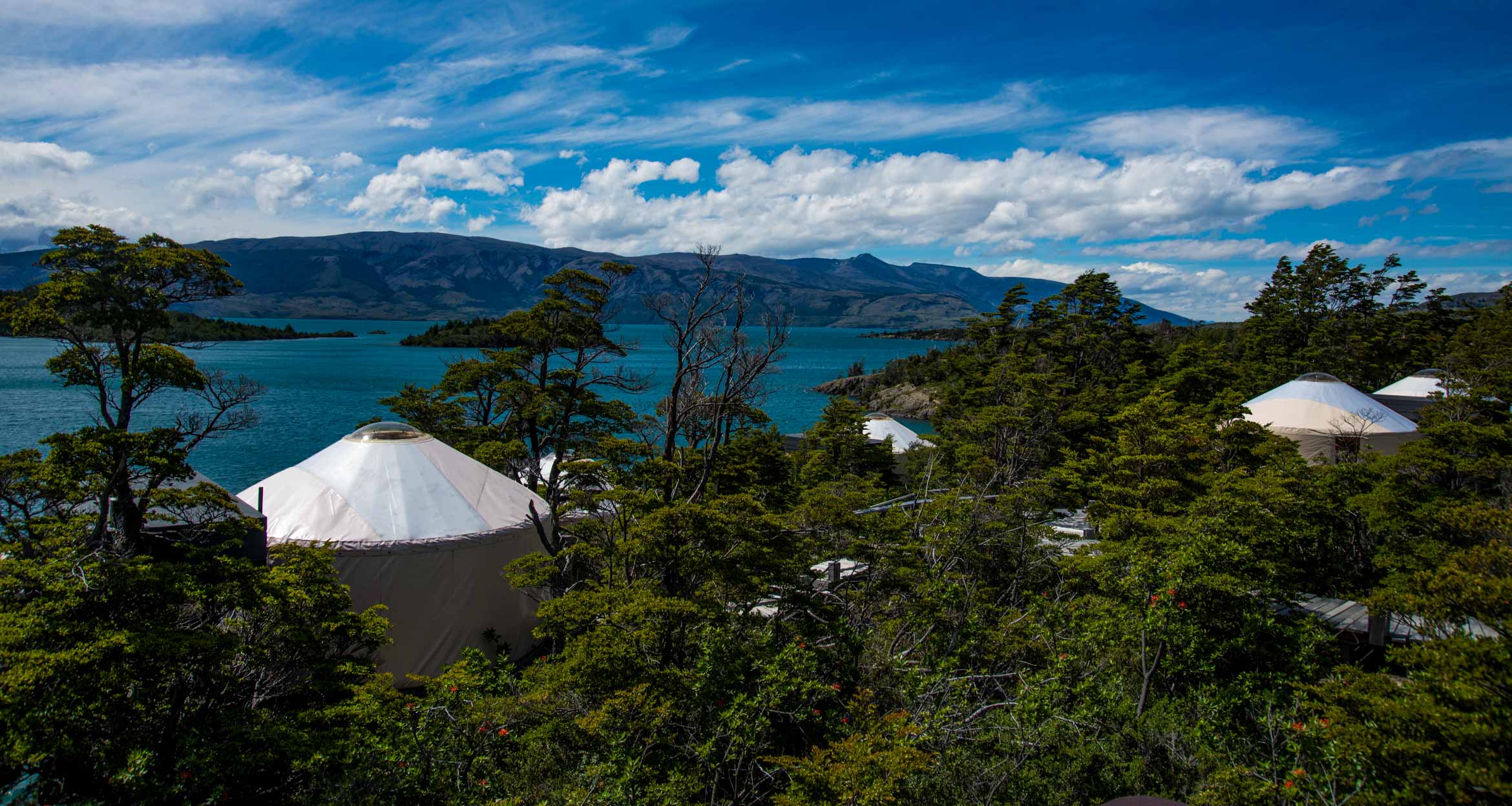 Yurts at Patagonia Camp, surrounded by lush green trees, overlook a blue lake and the distant Torres del Paine mountains under a bright, partly cloudy sky—luxury camping at its finest.