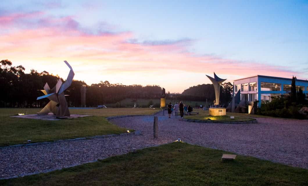 Modern sculptures and a group of people are seen in an outdoor park at sunset during the Coast to Coast Tour, with a contemporary building on the right and trees silhouetted against a colorful sky in the background.
