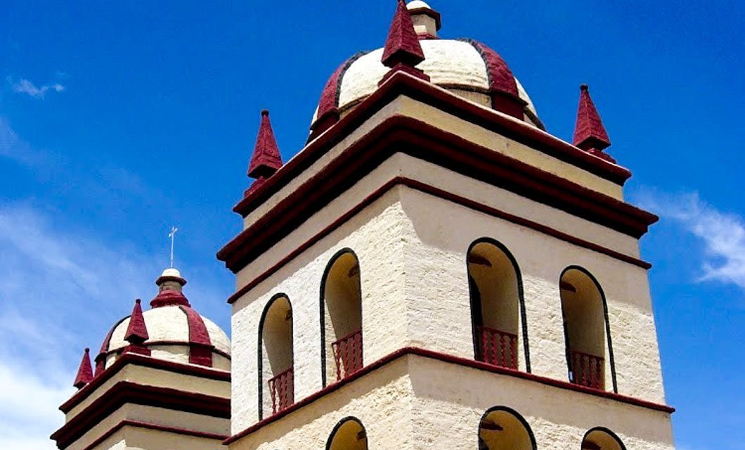 Two cream-colored, domed towers with red trim and pointed spires rise against a bright blue sky—an architectural gem you might spot on a scenic Peru Bolivia rail journey, featuring historic arches and ornate balconies.