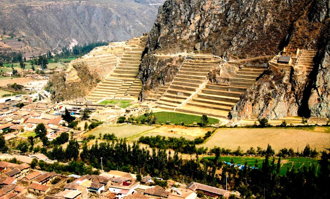 Terraced agricultural fields are carved into a steep mountainside near Machu Picchu, with a village of brown-roofed buildings and green patches below, all surrounded by majestic mountains in the background.