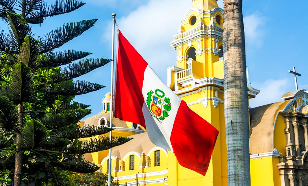 A Peruvian flag waves in front of a bright yellow colonial-style church with a bell tower and cross, capturing the essence of Peru against a blue sky and surrounded by palm and evergreen trees.